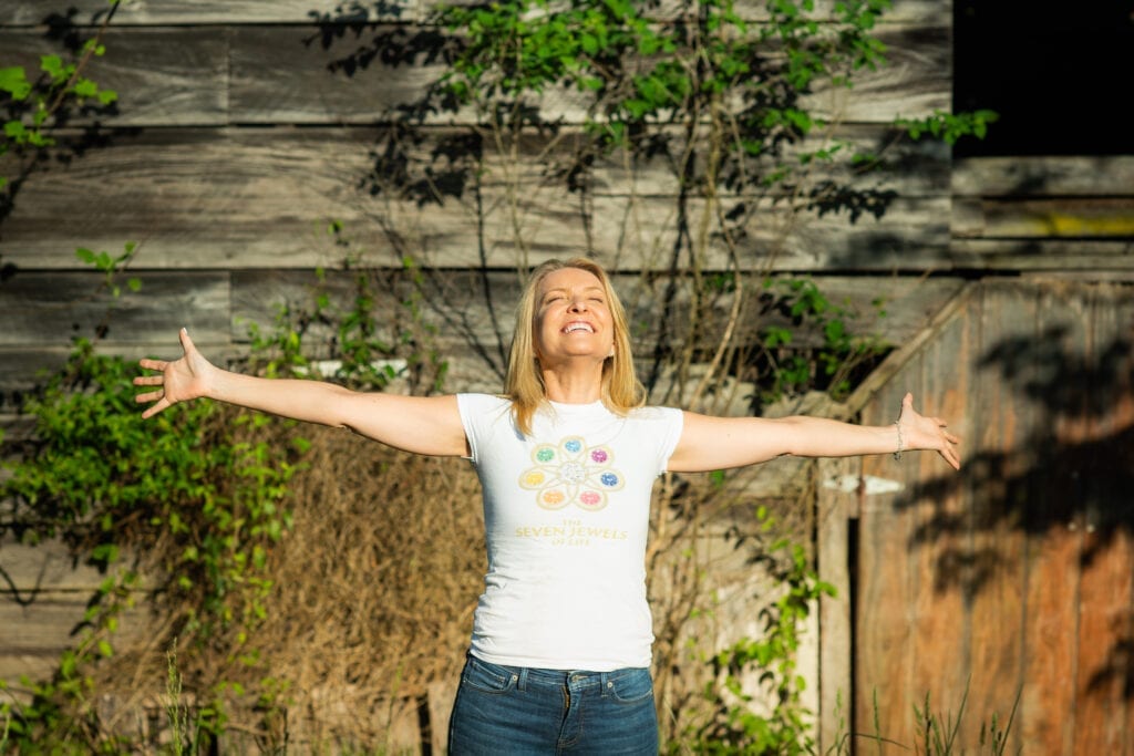 A blonde woman wearing a white t-shirt standing in a yard with her arms wide open